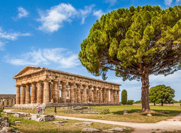 Tempio antico con colonne doriche e un grande albero, sotto un cielo azzurro.